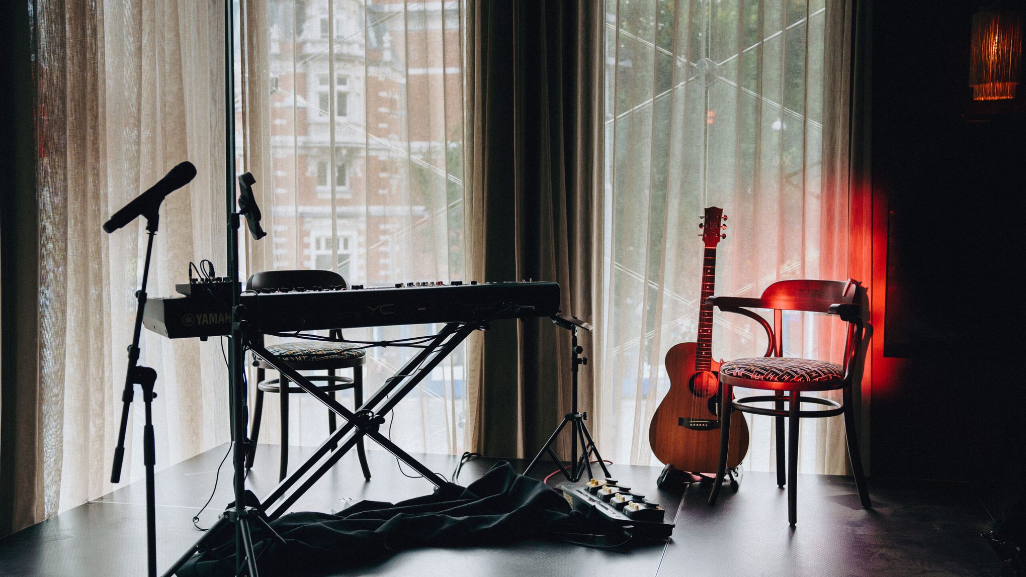 Small stage with a keyboard, microphone and acoustic guitar in front of tall curtained windows
