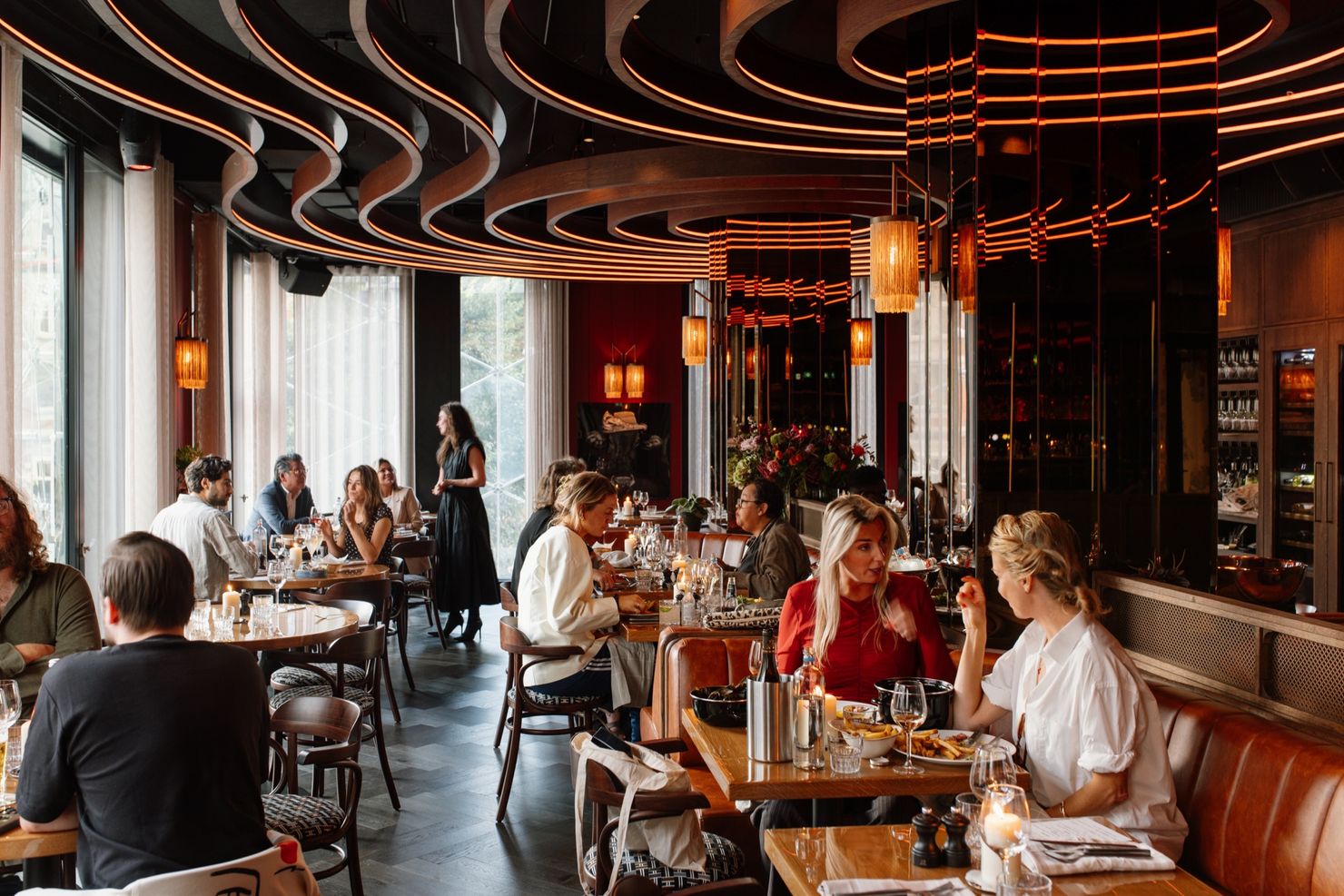 Guests at tables in the Bistro by daylight, with leather banquettes and tall windows onto Leidseplein