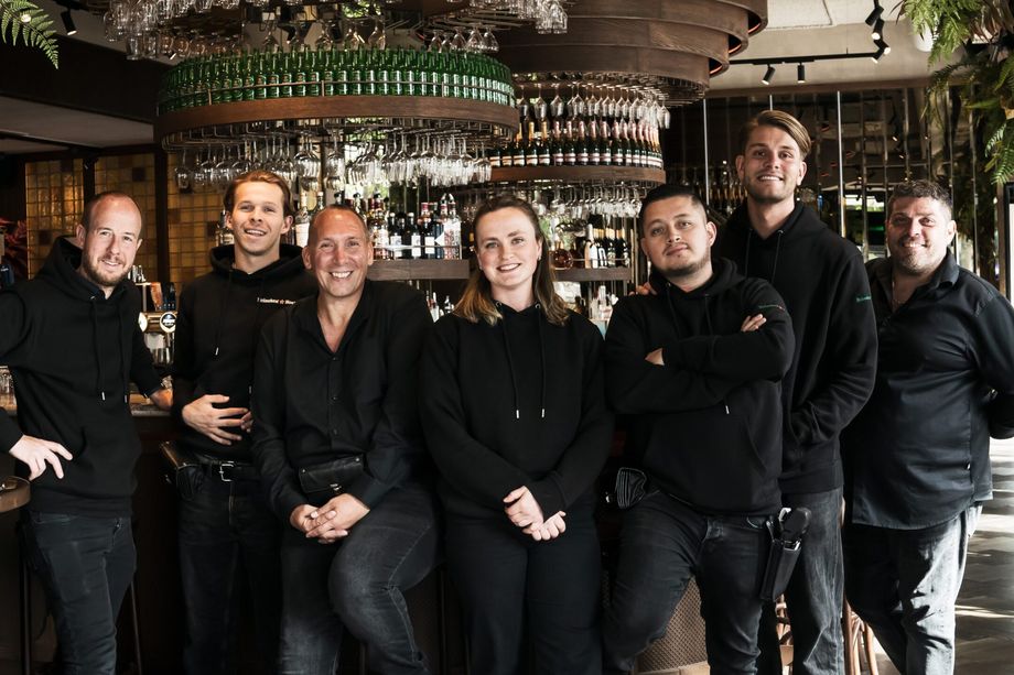 Het team van Heineken Hoek, zeven mensen in het zwart, lachend bij de bar van het Grand Café