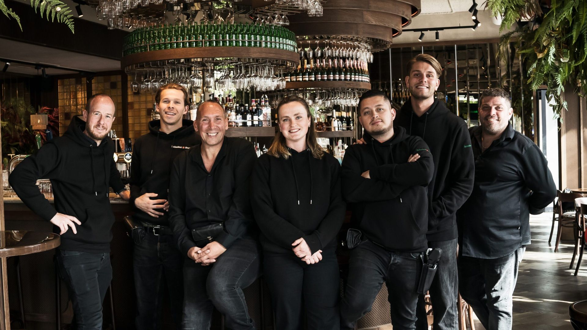 Het team van Heineken Hoek, zeven mensen in het zwart, lachend bij de bar van het Grand Café