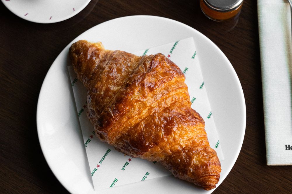 Een roombotercroissant op een servetje van Heineken Hoek op een wit bord, met een potje jam op een donkere houten tafel