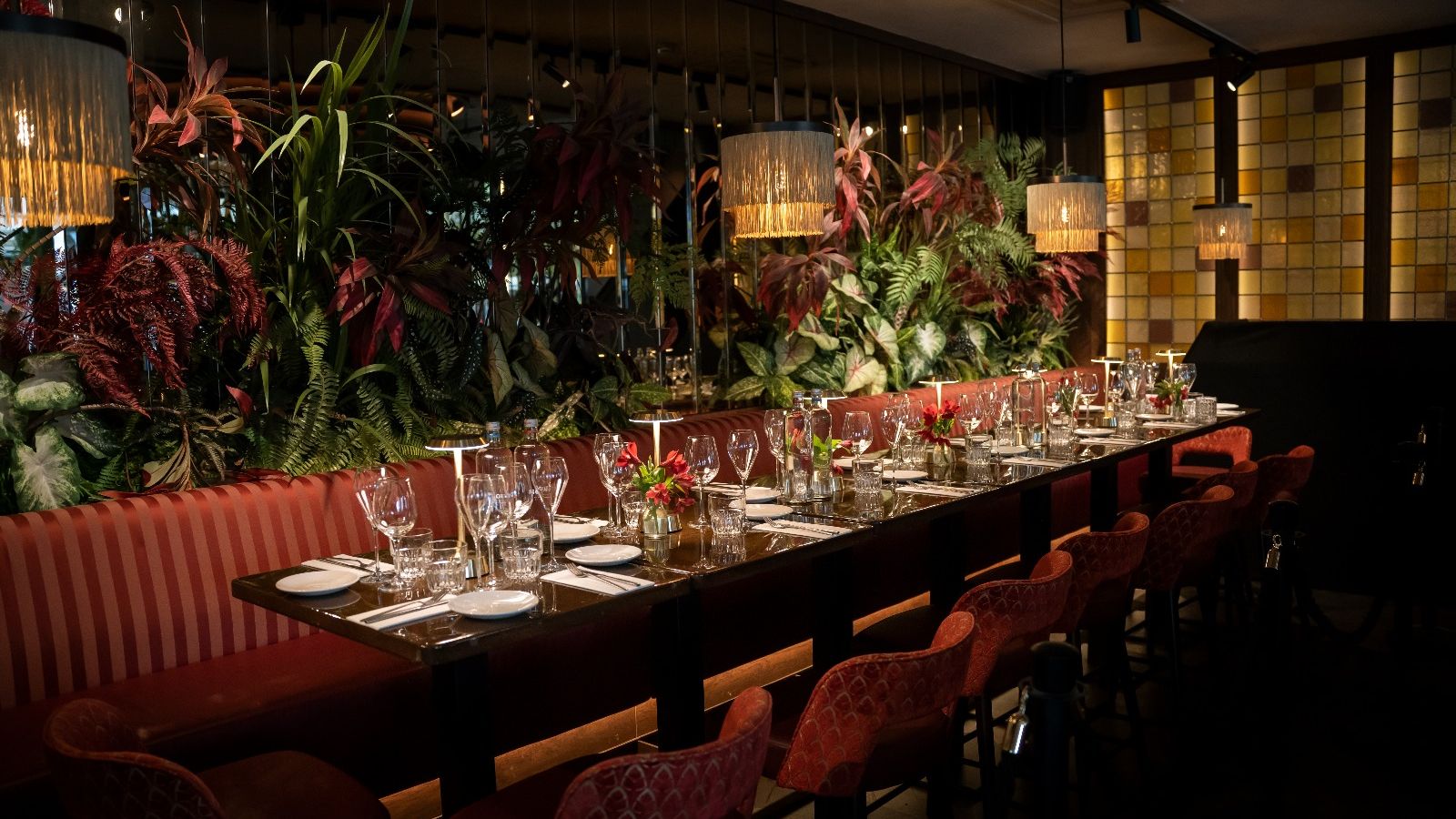 Een lange tafel gedekt voor een groepsdiner, met rode fluwelen stoelen, een gestreepte bank, planten en hanglampen met franje